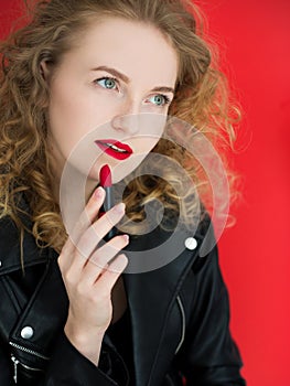 Portrait of young girl painting her lips with matte red lipstick on red background. Beauty, fashion concept.