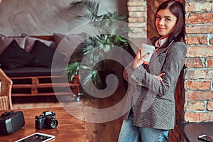 Portrait of a young girl photographer dressed in a gray elegant jacket holding cup of takeaway coffee while leaning on a