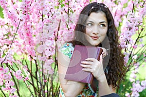 Portrait of young girl sitting near bush with pink flowers and reading the book