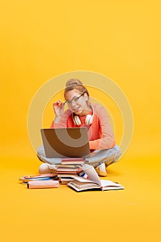 Portrait of young girl, student in glasses and sweater sitting on floor, studying isolated over yellow background