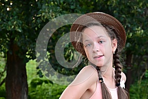 Portrait of young girl with two braids and hat, posing against background of green foliage.