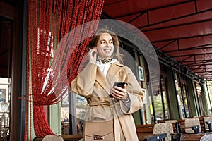 Portrait of young girl wearing coat listening to the music on cafe veranda with earphones holding cell phone in hands