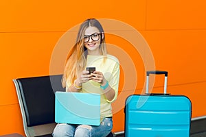 Portrait of young girl in yellow sweater sitting on chair on orange background. She has blue laptop on knees and blue