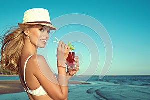 Portrait of young glam smiling blond lady in white swimming bra and panama drinking cocktail through a straw