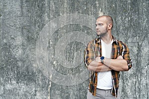 Portrait of young good looking bold bearded guy standing outdoors against grey loft wall looking aside with his hands crossed.