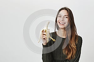 Portrait of young gorgeous brunette model smiling sincerely, holding banana over white background. Healthy lifestyle and