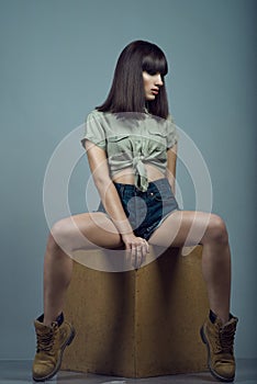 Portrait of young gorgeous dark-haired model wearing high waisted dark blue jeans shorts, khaki shirt and boots sitting on cube