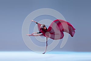 Portrait of young graceful ballerina dancing with deep red transparent fabric isolated over blue grey studio background