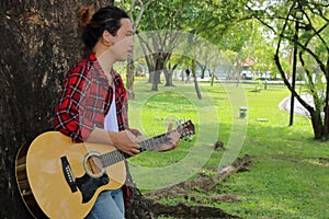 Portrait of young guitarist standing against a tree and playing acoustic guitar in beautiful nature background.