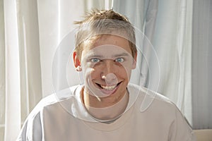 Portrait of a young guy 20-25 years old, sitting by the window and smiling. Concept: good news, good mood.