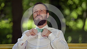 Portrait of a young guy with a beard who sits on a bench in the park and adjusts his bow tie.