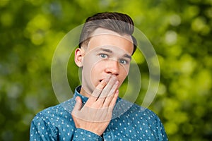 Portrait young guy dressed blue shirt. man open his eyes wide with hands, fingers and mouth. Man on green bokeh background