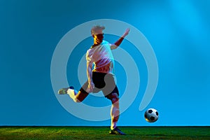 Portrait with young guy, man football soccer player kicking ball in action over soccer field background in neon light