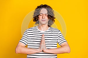 Portrait of young guy wearing stylish striped t shirt clasped hands dreaming and practicing yoga isolated on yellow
