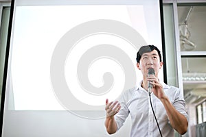 Portrait of young handsome Asian male speaker publicly speaking on stage to group of audience with white board behind.