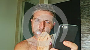 Portrait of young handsome and attractive internet addict man at home bathroom with towel on shoulder washing teeth with