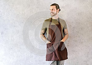 Portrait of young handsome barista man in apron with tattooed arms against the background of a gray wall in a loft cafe and