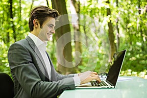 Portrait of young handsome business man in suit at laptop at office table in green forest park. Business concept.