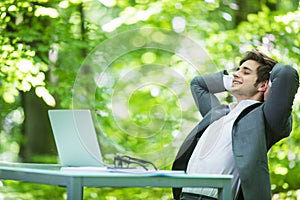 Portrait of young handsome business man in suit relax at laptop at office table hands over head in green forest park. Business con