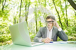 Portrait of young handsome business man in suit working at laptop at office table in green forest park. Business concept.