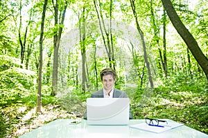 Portrait of young handsome business man in suit working at laptop at office table in green forest park. Business concept.