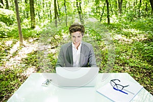 Portrait of young handsome business man in suit working at laptop at office table in green forest park. Business concept.