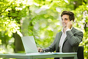 Portrait of young handsome business man in suit working at laptop at office table in green forest park. Business concept.