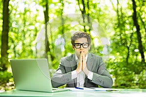 Portrait of young handsome business man in suit working at laptop at office table with pray hands in green forest park. Business c