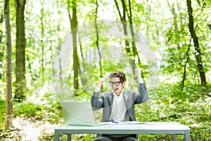 Portrait of young handsome business man in suit working at laptop at office table with raised hands achived goals in green forest