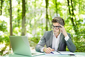 Portrait of young handsome business man in suit working at laptop at office table thinking in green forest park. Business concept.