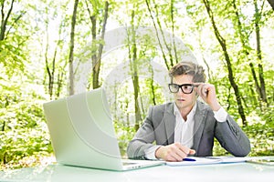 Portrait of young handsome business man in suit working at laptop at office table thinking in green forest park. Business concept.