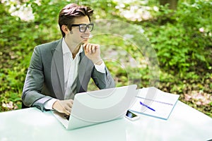 Portrait of young handsome business man working at laptop at office table with hand on chin smile in green forest. Business concep