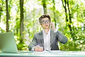 Portrait of young handsome business man working at laptop at office table and talk at phone with costumer and make notice in green