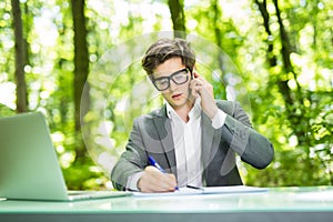 Portrait of young handsome business man working at laptop at office table and talk at phone with costumer and make notice in green