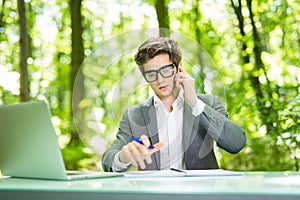 Portrait of young handsome business man working at laptop at office table and talk at phone with costumer and make notice in green