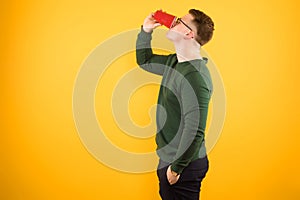 Portrait of young handsome confident guy in green sweater drinking from red plastic cup on yellow background