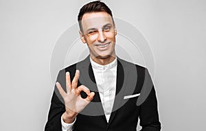 Portrait of a young handsome happy man, in a business suit, smiling, and showing an OK gesture, on gray background.