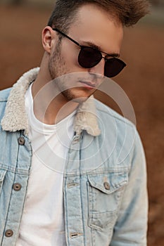 Portrait of a young handsome man with a beard with a fashionable hairstyle in stylish sunglasses in a white t-shirt in a denim