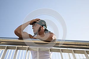 Portrait of a young handsome man dressed in jeans and white t-shirt and green headscarf. The young man is leaning on railing and