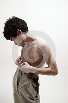 Portrait of young handsome man with fit body, posing shirtless in beige pants against white studio background