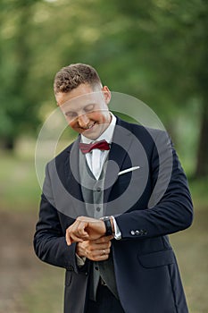 Portrait of young handsome man groom wearing blue suit, grey vest, red bow tie, looking at watch in park in summer.