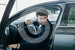 Portrait of young handsome man in his new silver gray car, relaxing, hand on steering wheel, looking out window,  on