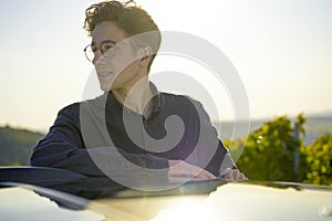 Portrait of a young and handsome man leaning at his car looking aside with a beautiful greenery in the background