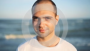 Portrait of young handsome man looking and smiling at camera standing on the beach with a sea on background. Happy