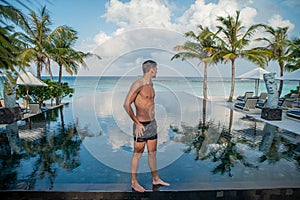 Portrait of young handsome man model standing on on the edge of swimming pool at the tropical island luxury resort