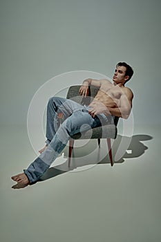 Portrait of young handsome man with muscular relief body sitting on chair, posing shirtless in pants over pale green