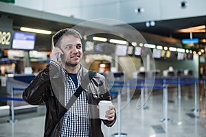 Portrait of young handsome man walking in modern airport terminal, talking smart phone, travelling with bag and coffee