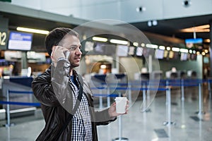 Portrait of young handsome man walking in modern airport terminal, talking smart phone, travelling with bag and coffee