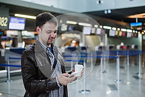 Portrait of young handsome man walking in modern airport terminal, texting, travelling with bag and coffee, wearing