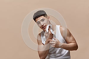 Portrait of young handsome man wiping face after shaving, taking care after skin against light brown studio background